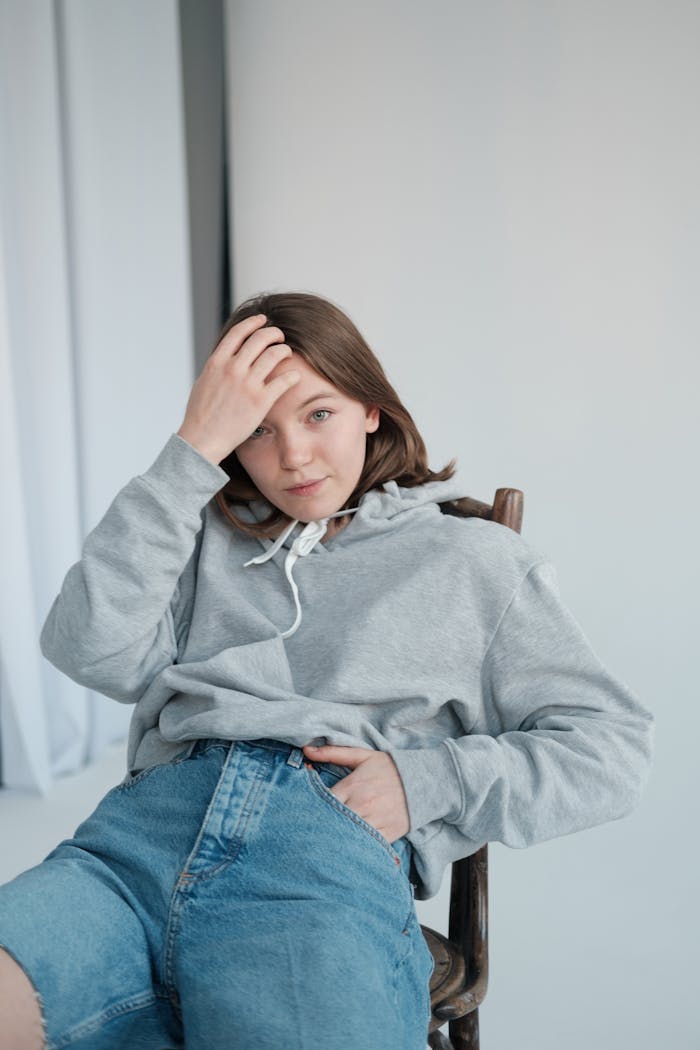 Serious female model wearing sweatshirt and denim shorts attaching hair while sitting on chair and looking at camera